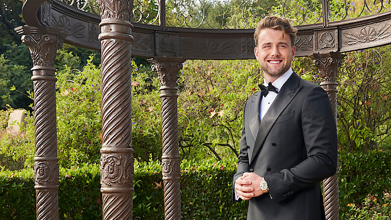 Man in a black tuxedo smiling outdoors, standing near ornate pillars and greenery, with an elegant garden gazebo structure in the background.