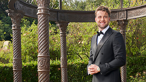 Man in a black tuxedo smiling outdoors, standing near ornate pillars and greenery, with an elegant garden gazebo structure in the background.
