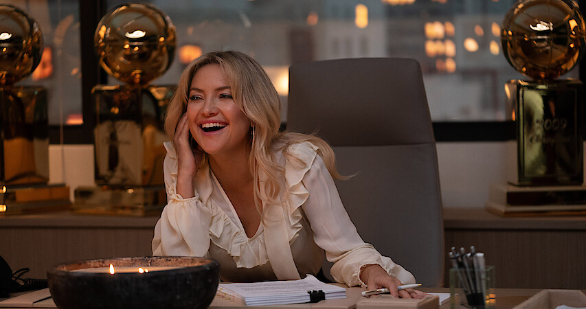 Kate Hudson as Isla Gordon sits at a desk smiling surrounded by basketball trophies in season 1 of 'Running Point'