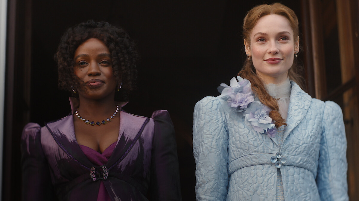 Duas mulheres vestidas com trajes de época elaborados, uma de roxo e outra de azul-claro com detalhes florais, estão lado a lado diante de um fundo escuro, olhando para a frente com expressões serenas, em um cenário histórico.