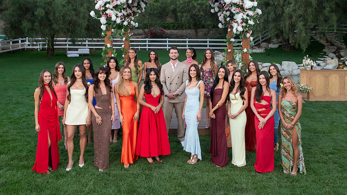 A group of women in colorful dresses and one man in a beige suit stand outdoors on grass, under floral arches, surrounded by trees and wooden fences, in a formal garden setting.
