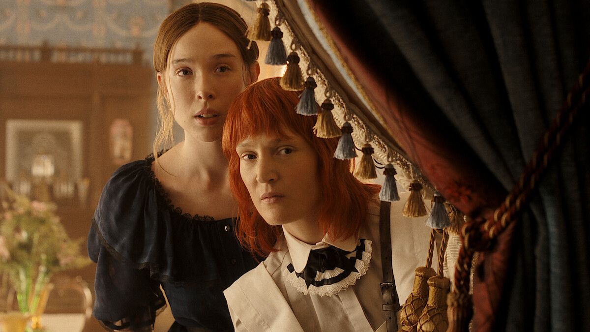 Two women in vintage clothing stand closely together, peering out from behind ornate curtains in an elegant, softly lit room with classic decor and floral arrangements, suggesting a historical or period drama setting.
