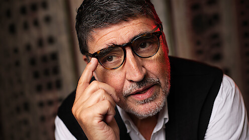 Alfred Molina with his hand raised to his glasses wearing a sweater vest and white button up shirt.