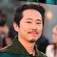 Steven Yeun, com cabelo escuro curto e cavanhaque, veste uma camisa verde e posa ao ar livre em um evento com uma multidão desfocada e iluminação aconchegante ao fundo.