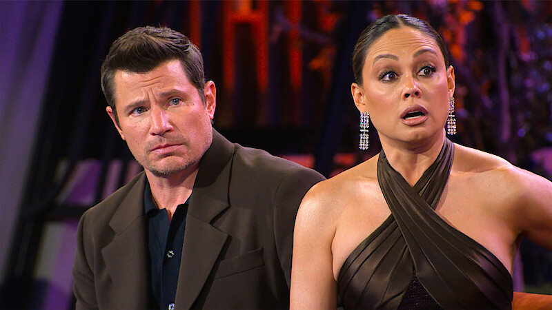Nick and Vanessa Lachey dressed formally sit indoors, reacting with surprised and serious facial expressions, with colorful lights and abstract decor in the background.