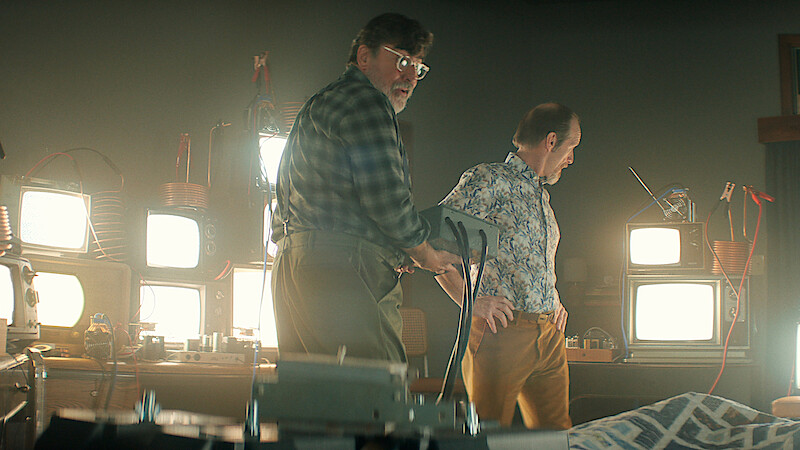 Two men in a dimly lit room filled with old televisions and electronic equipment, one holding a device, standing beside a bed with a patterned blanket, suggesting an experimental or scientific setting.