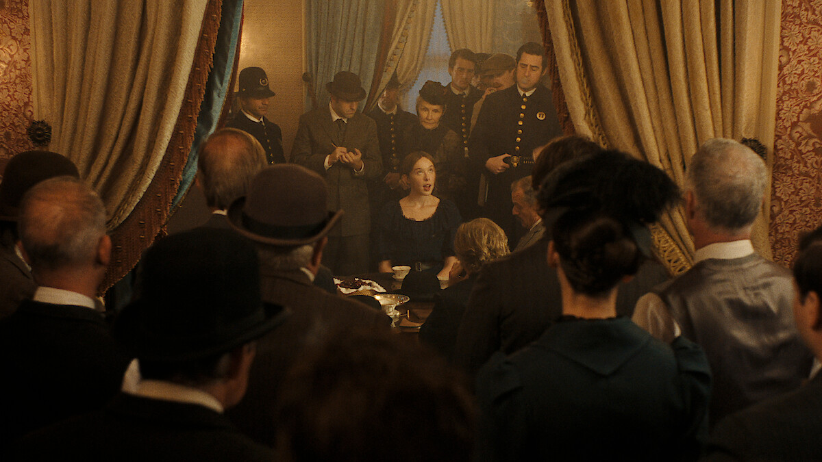 A woman sits at a table in a vintage, ornately decorated room, surrounded by police officers and people in period clothing, all focused on her as if witnessing an important event or announcement.