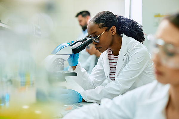 Woman wearing a lab coat looking through a microscope in a lab environment.
