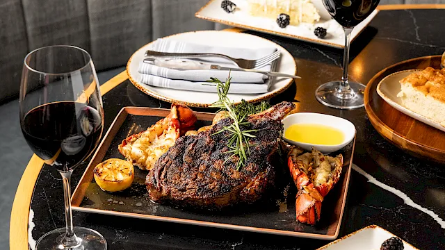 A fine-dining plate with grilled steak, lobster, lemon, herbs, two glasses of red wine, and cheesecake topped with berries.