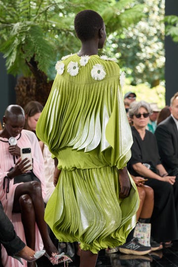 Model in green pleated dress with white floral accents at fashion show.