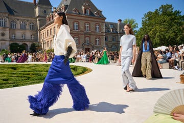 Models walking on outdoor fashion runway with audience and historic building in background.