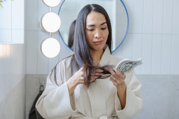 Woman in bathrobe examining hair with a brush in front of a bathroom mirror during morning routine