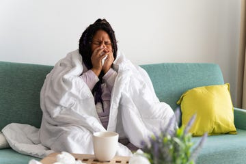 common pneumonia symptoms - young woman sitting on the sofa at home, covered with a blanket, blowing her nose, suffering from a cold or flu
