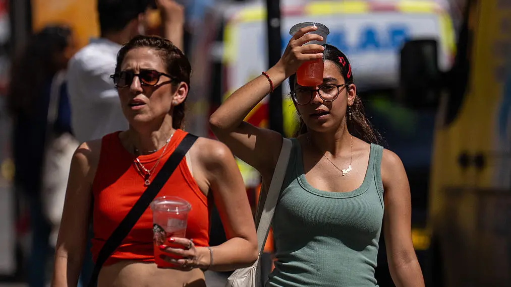 Two women walking along holding cold drinks, as one of them holds it to her forehead to cool down in the warm weather