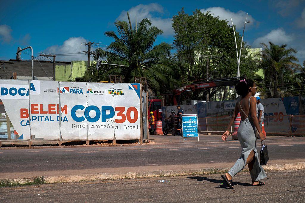 A woman walks past an infrastructure project under way for COP30 in Belem. She has her hair up and is wearing a grey dress and carrying a black bag. Misaligned posters in white, blue and orange read "Belem Capital Da COP30".