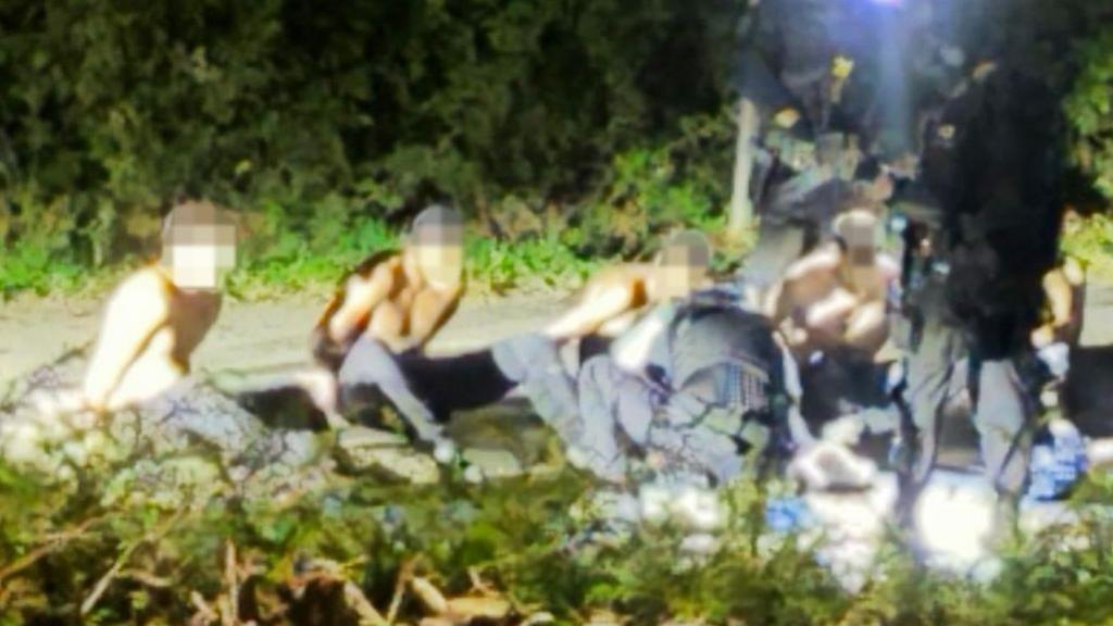Detained men in Whelford. Their faces are blurred. They are shirtless. Trees line the background.