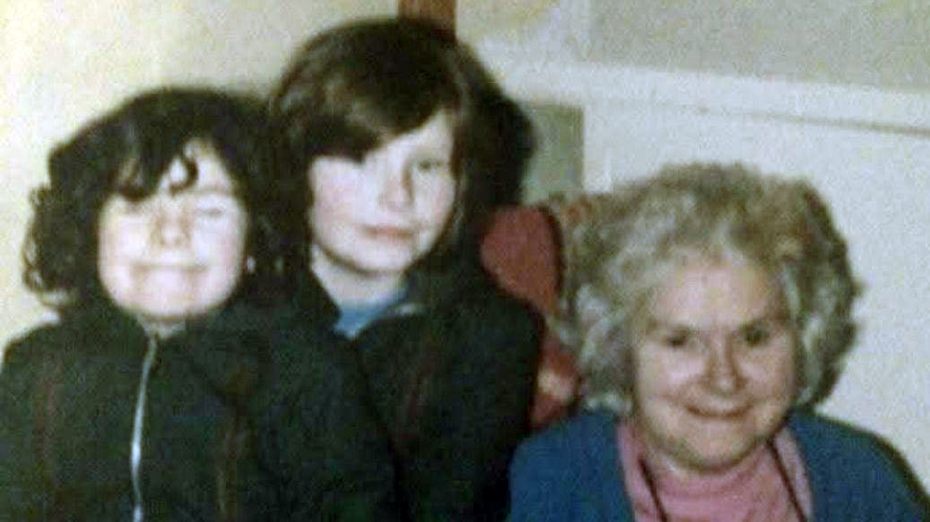 An old photograph of two young girls with dark hair and their grandmother. The girls are standing together next to an older woman who's sitting in a chair. They're all looking at the camera. 