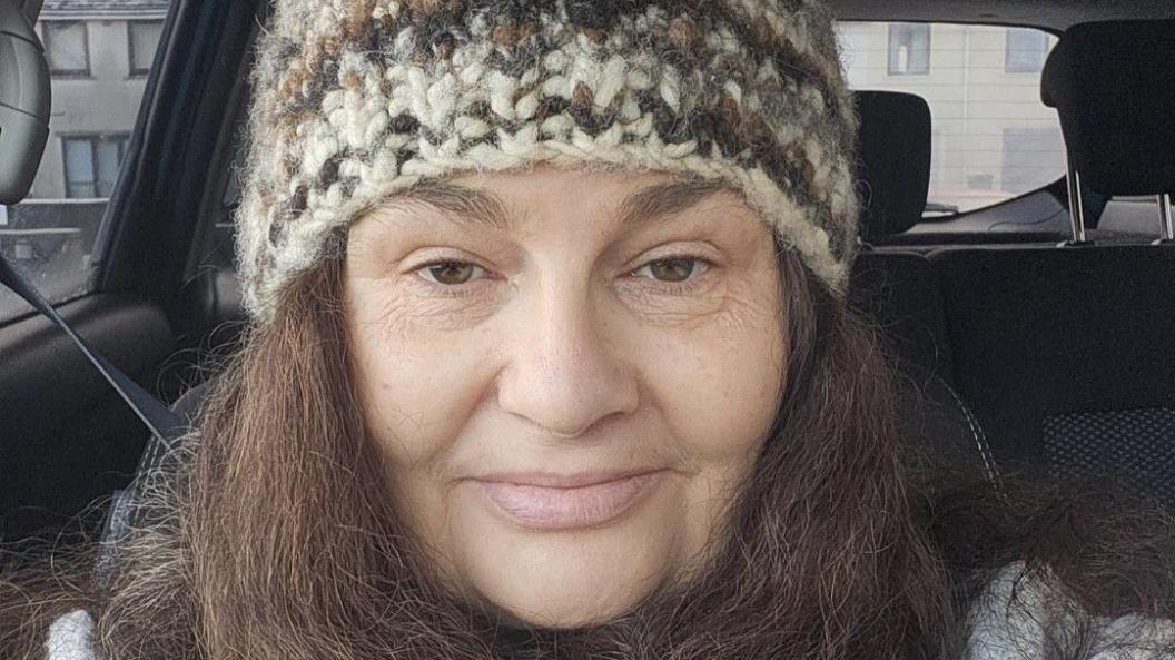 Diane Barnes is a middle-aged woman. She is sitting in a car and taking a selfie. She has dark hair and is wearing a knitted hat. There are houses in the distance. 