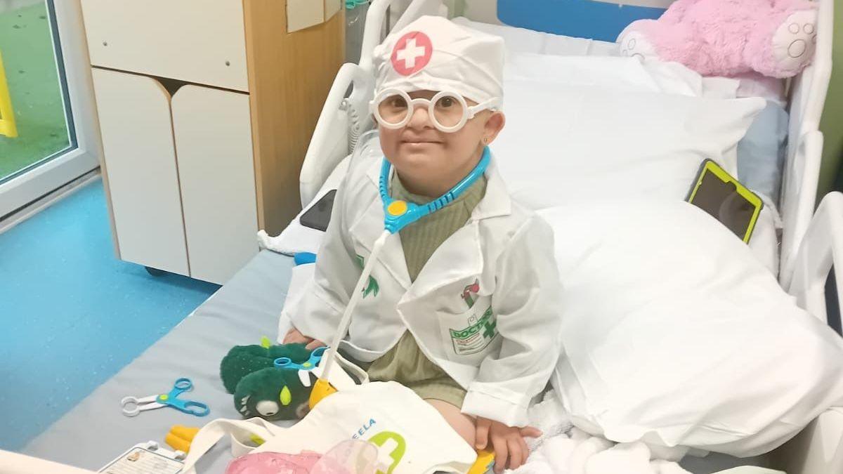 A young girl is sitting on a hospital bed and she is dressed up in a doctor's uniform including white scrubs and a stethoscope.