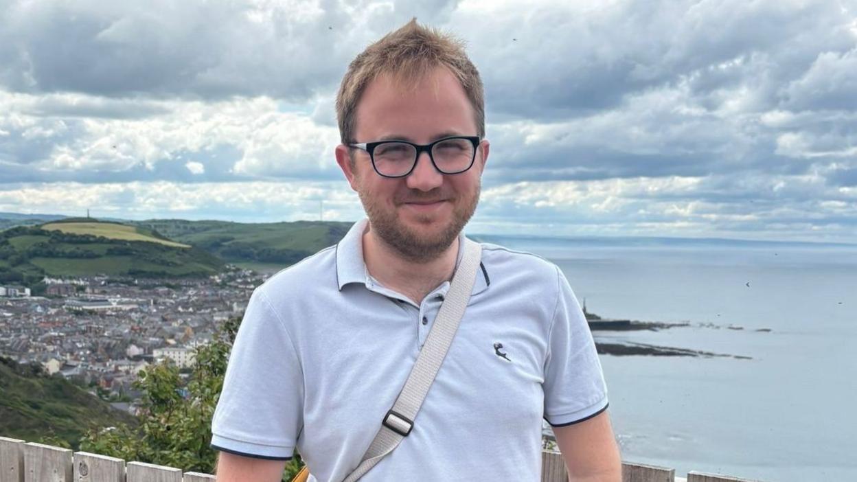 Nick smiling at the camera on coastline. He is wearing a white polo shirt, glasses and has a bag over his shoulder.