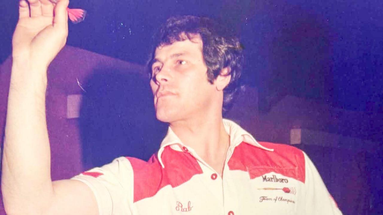 Rab Smith has dark hair and is wearing a white darts shirt with red shoulders as he throws a red dart