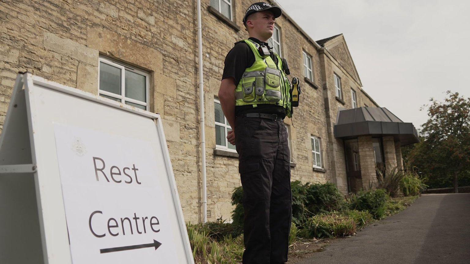 A policeman is standing next to a white A-frame sign, which says "rest centre" and has an arrow pointing right beneath the words. Behind the policeman are terraced houses built with light brown stone.