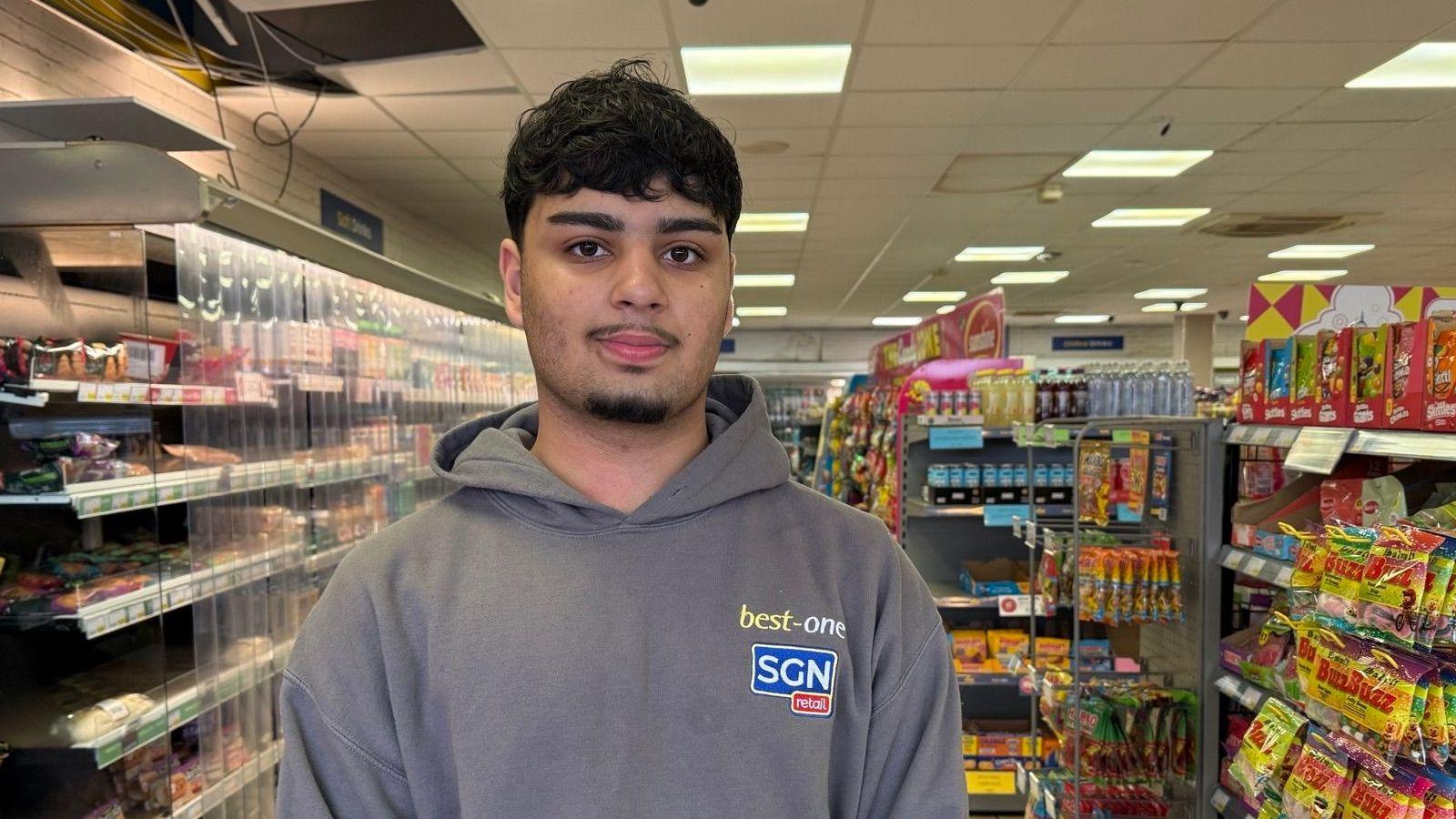 Hakim has black curly hair and wears a grey hoody with best-one and SGN retail embroidered on it. He stands in the shop with the aisles behind him. 