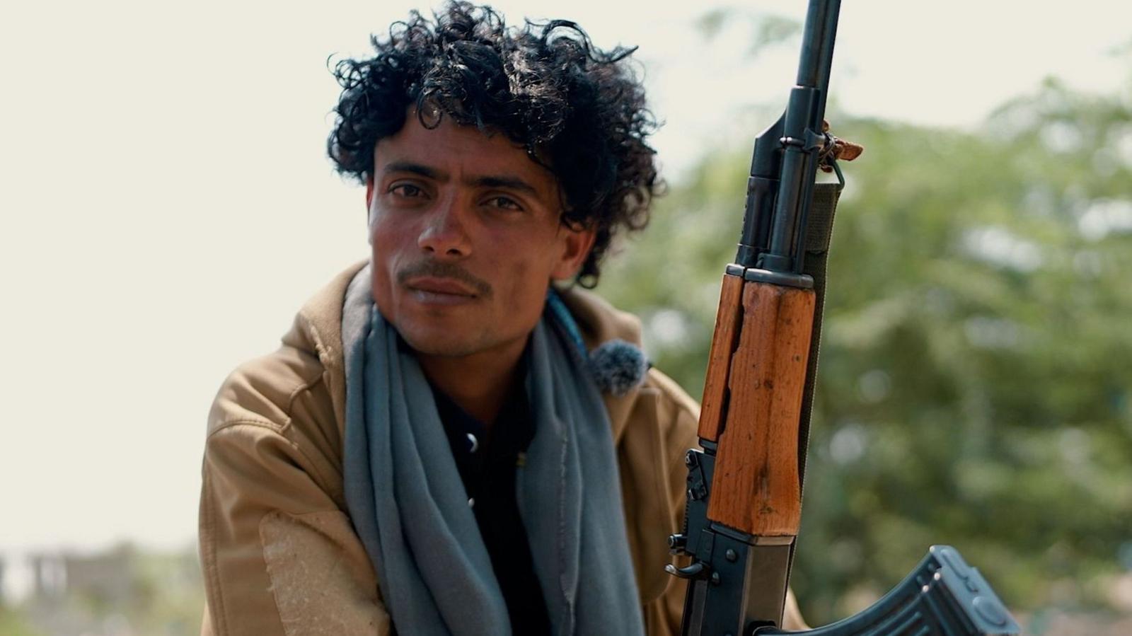 A man with curly hair holds a rifle
