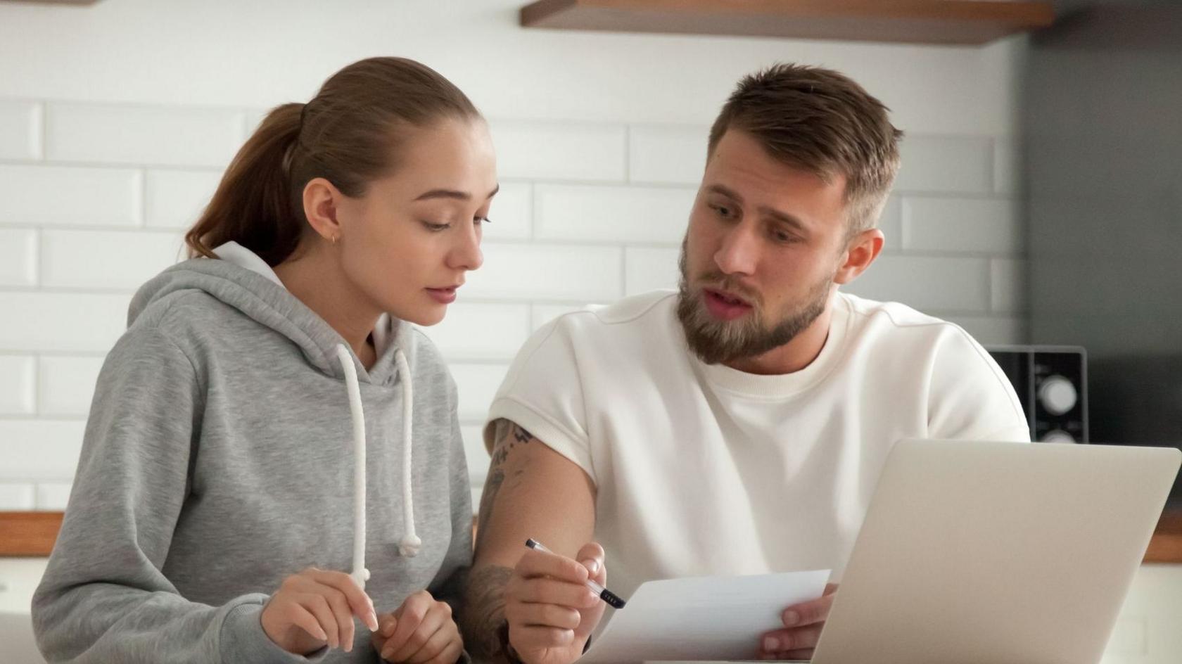 Couple look at a bill in their kitchen with a laptop in front of them.