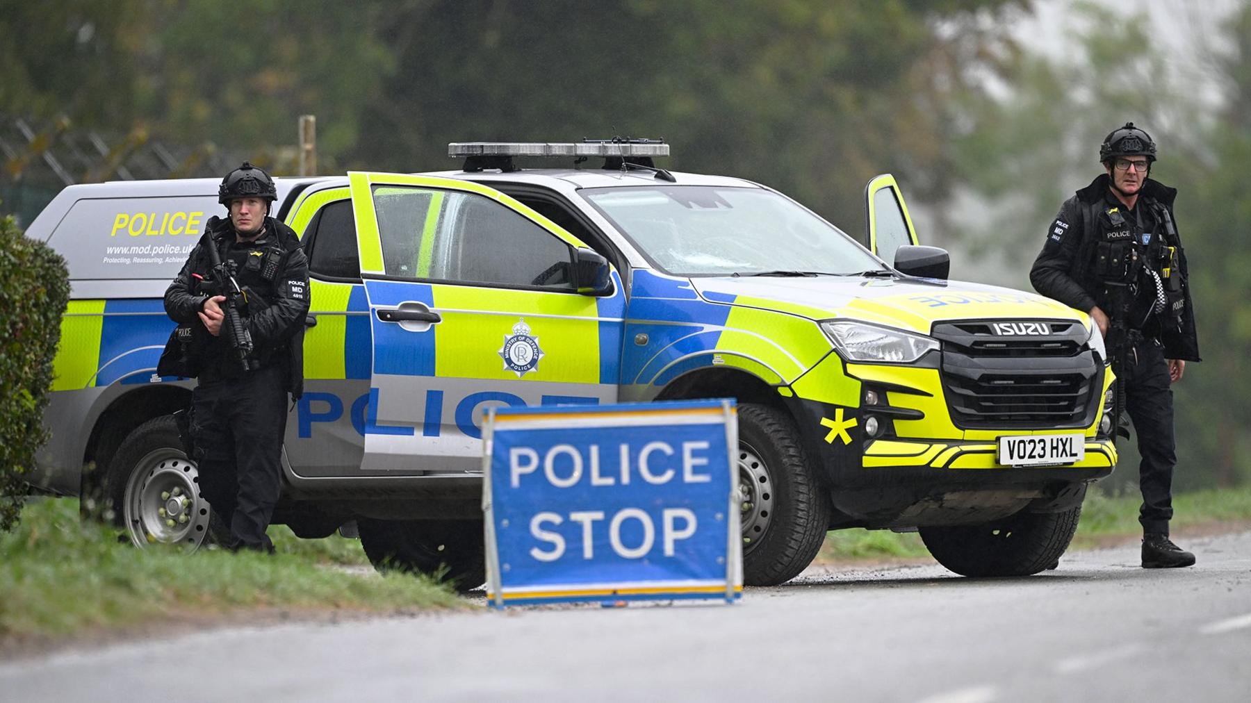 Two armed police men at Fairford  standing next to a yellow police van with a blue sign saying 'police stop' on the road in front of them