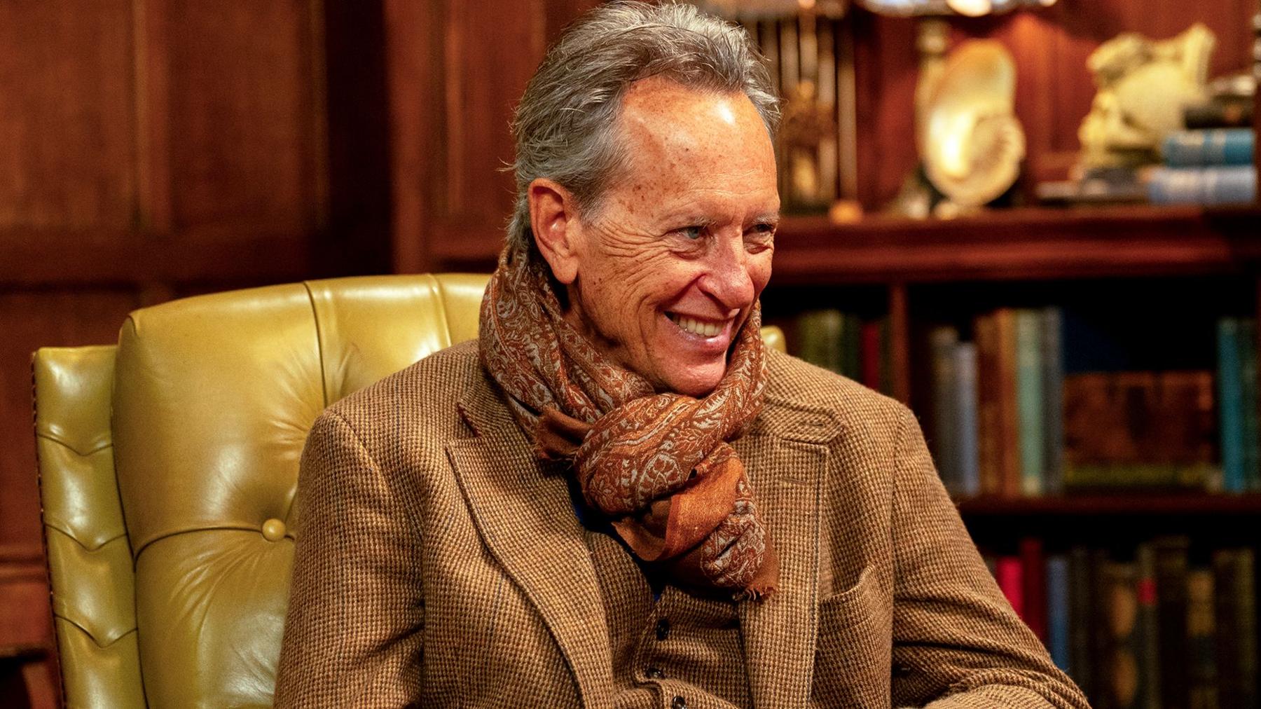 Richard E Grant wearing a tweed suit and scarf sitting on a leather chair and smiling 