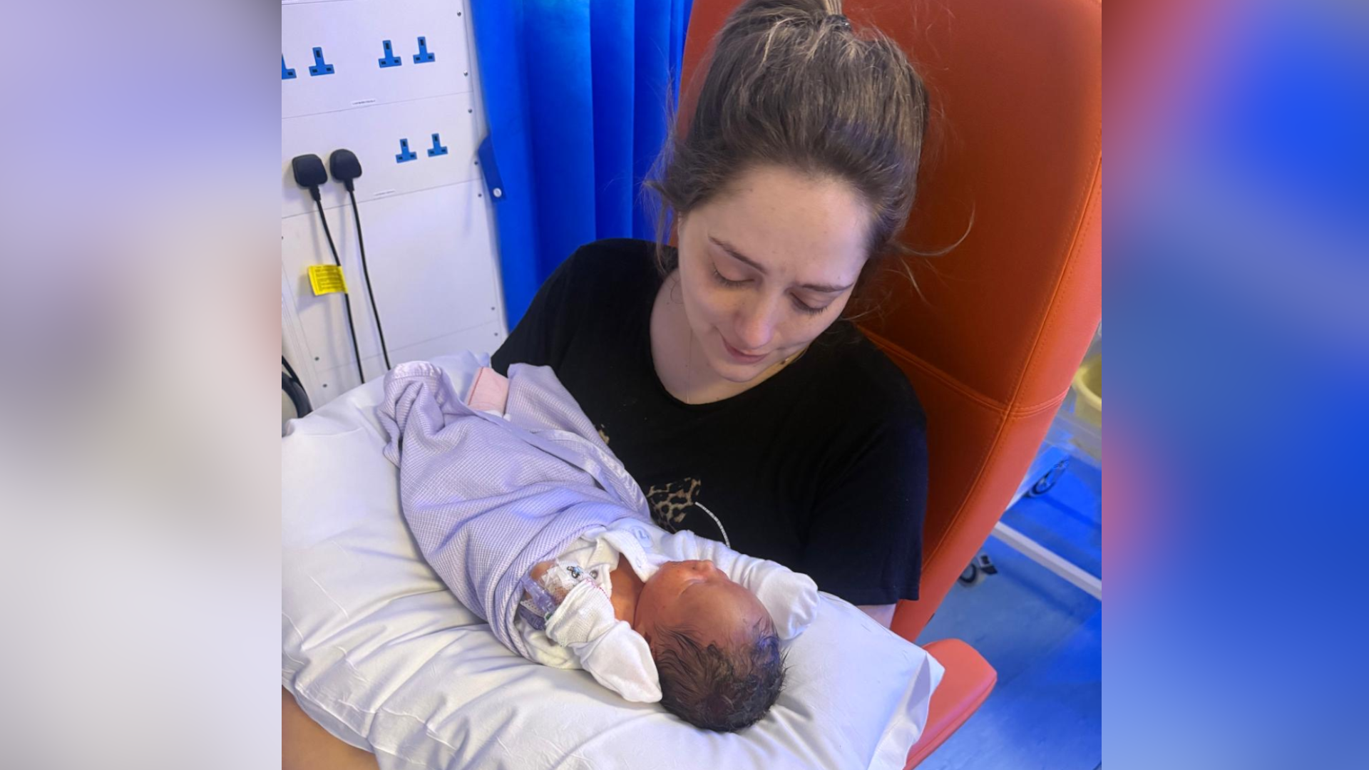 Ciara, a woman with brunette hair pulled back into a ponytail. She is wearing a black T-shirt and is not looking at the camera. Instead, she is looking at her baby which is laid on a pillow on her lap. The baby is in a baby-grow, wearing white mittens and is wrapped in a blanket.