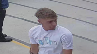 A CCTV image of a man with short, neat, light brown hair, looking to the side. He is wearing a white T-shirt with a hard-to-read logo printed across the chest. He appears to be stood outside during the daytime.
