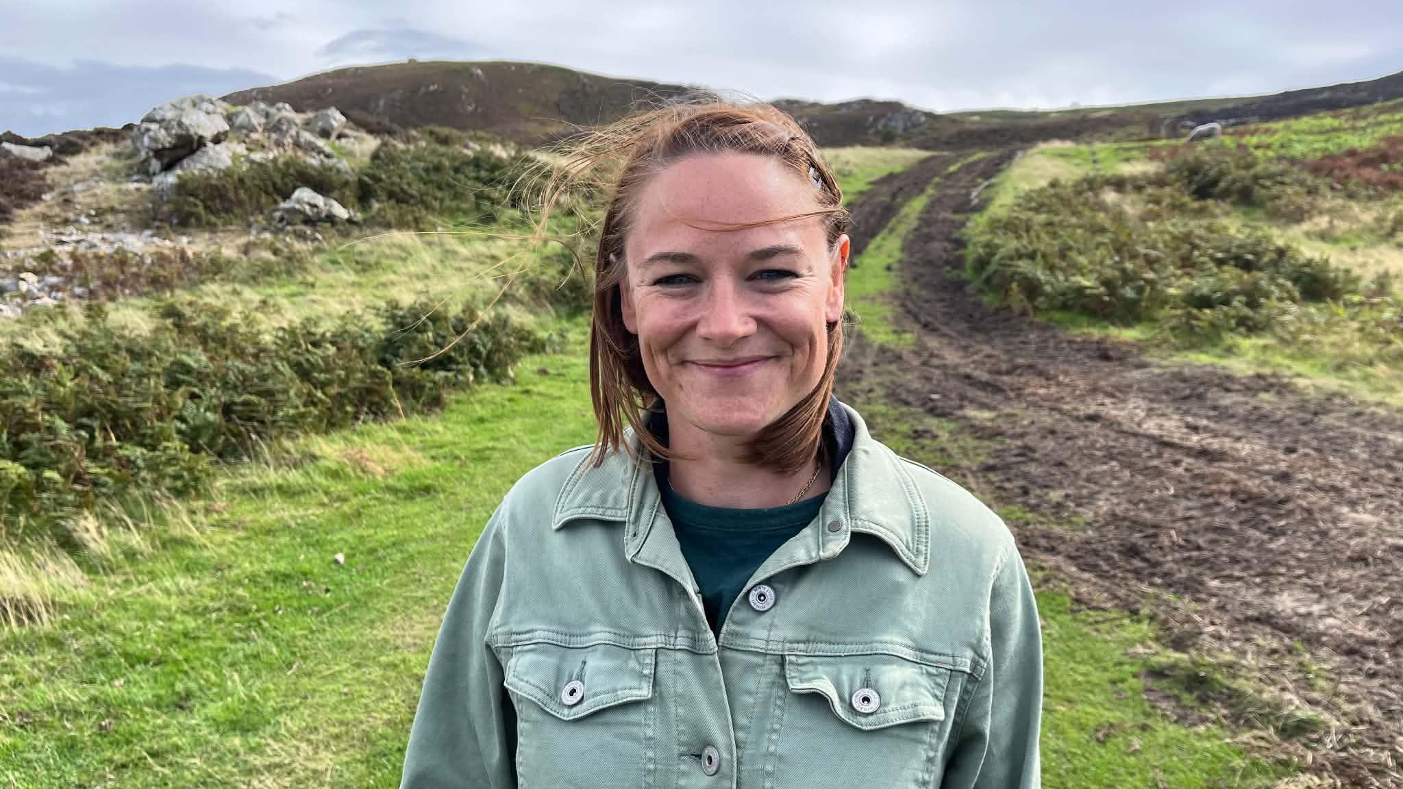 Kara wears a green denim jacket. She has short brown hair and stands in front of grassland and charred earth