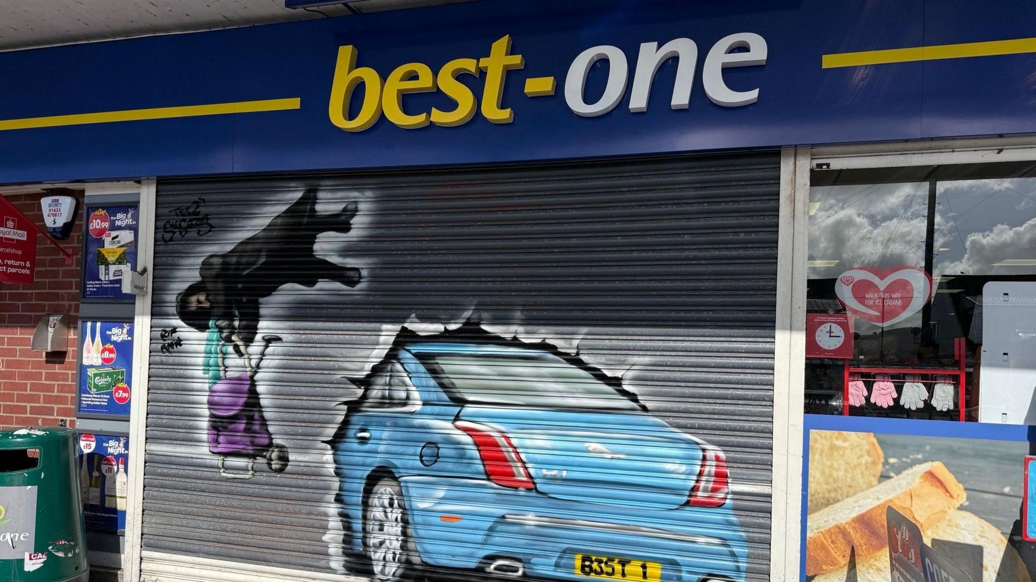 Graffiti artwork painted on a shop shutter showing a lady in a coat and scarf dangling in the air holding on to her trolley and a painting of a blue Jaguar smashing into the shop. 