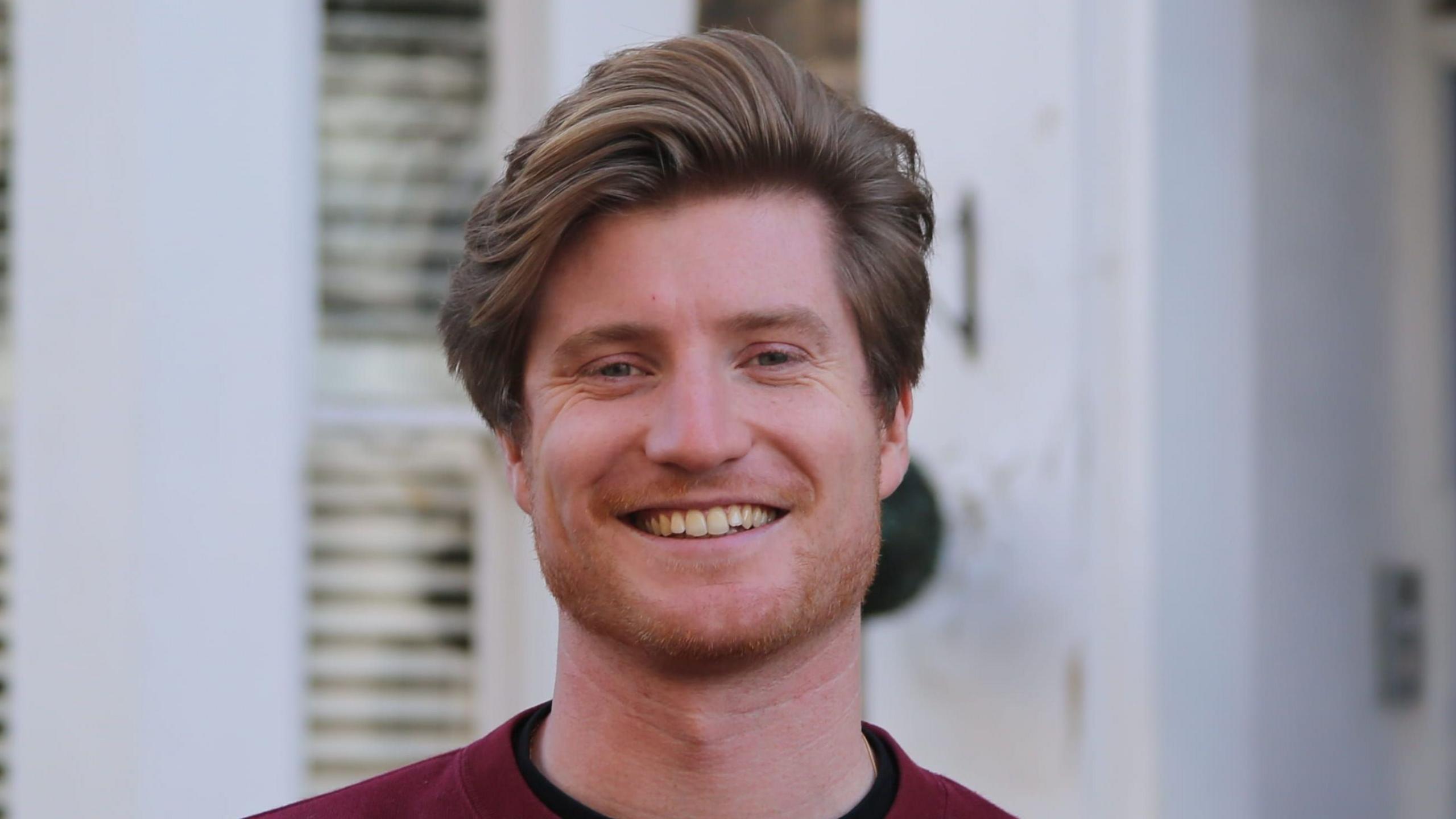Close-up portrait of Patrick Stillman seen a with thick, side-parted light brown hair and light stubble, wearing a dark red jumper outdoors with a blurred white building and door in the background.