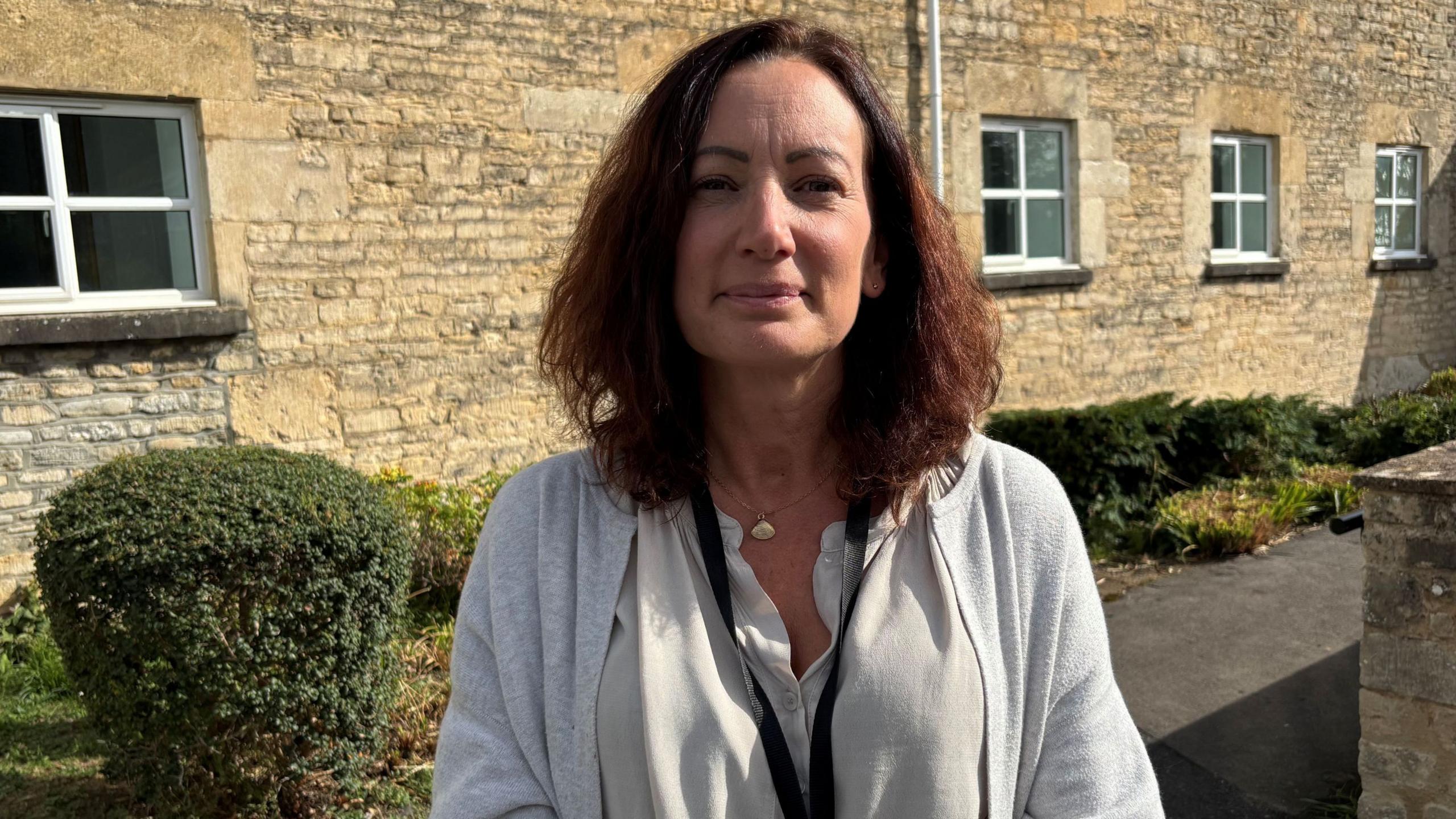 A woman with short dark hair is looking at the camera. She is wearing a grey top and a grey cardigan. Behind her is some bushes and a light brown stone building.