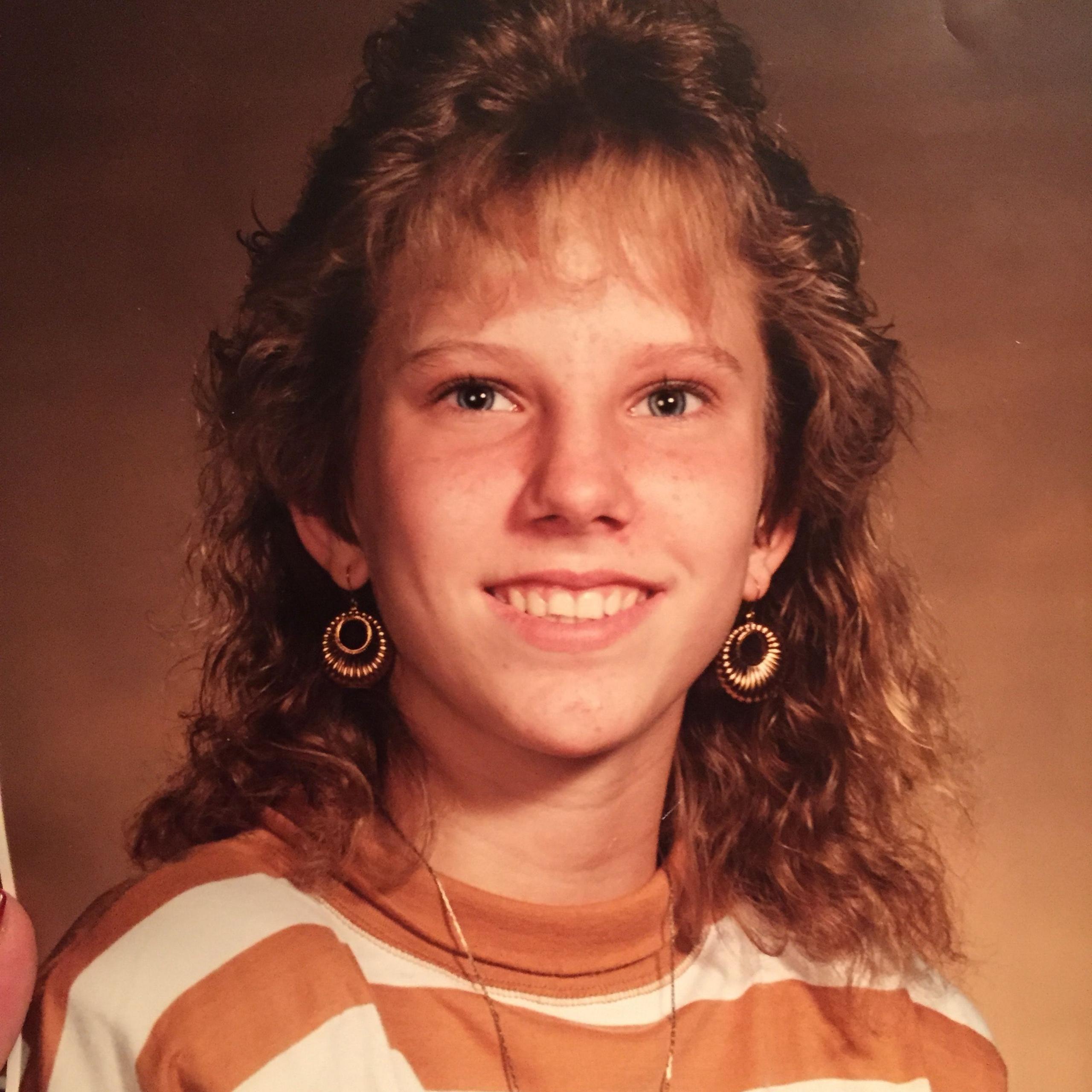 A portrait of Colleen Slemmer, taken in what looks like a photography studio with a backdrop. She is wearing a striped orange and white sweatshirt, gold drop earrings and is smiling at the camera. She has auburn hair and a fringe.