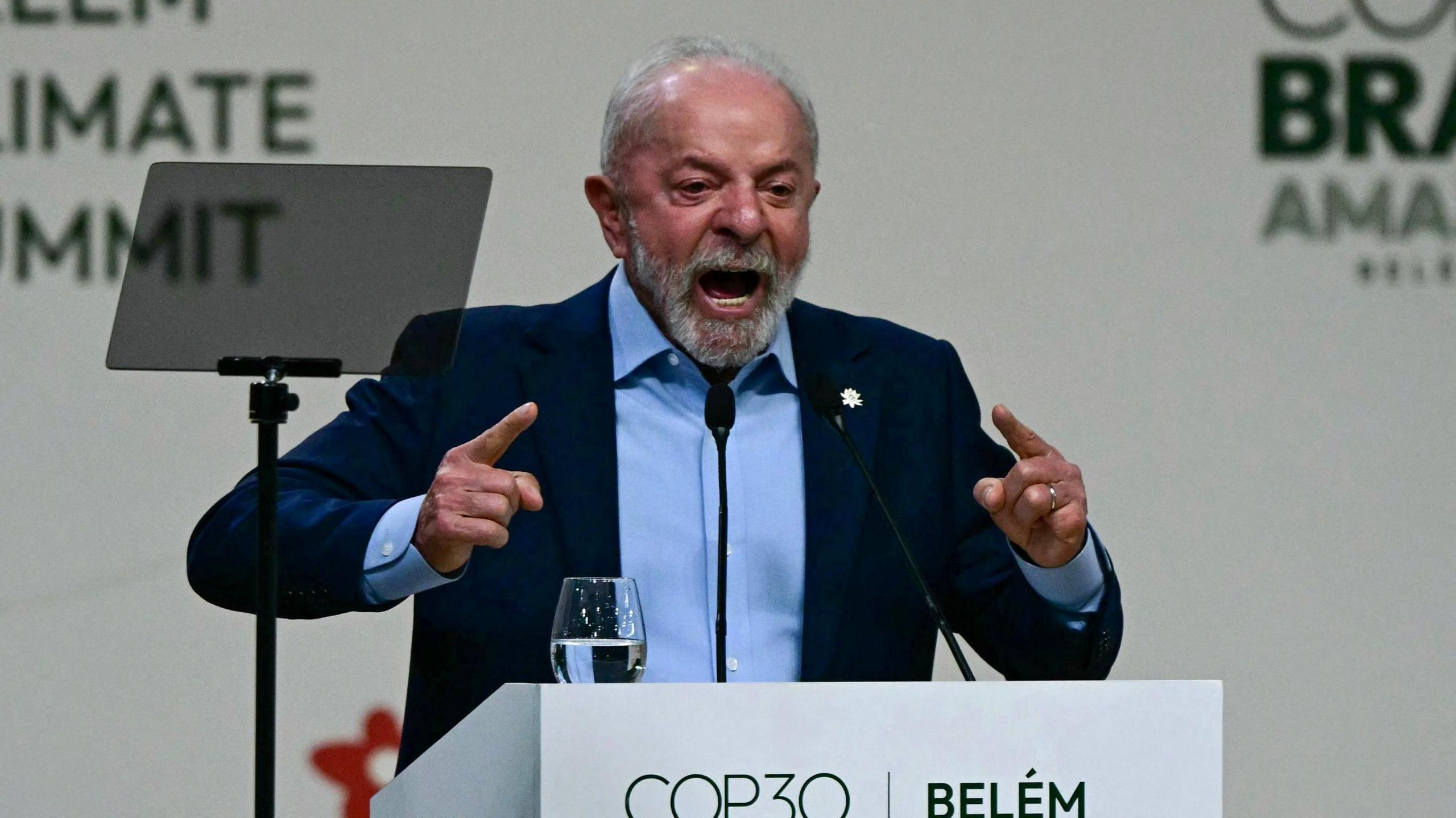Brazilian President Lula stands behind a white podium with the words "Belém COP30". On the podium is a glass of water. Lula is in a blue suit his hands pointing forward and his mouth open mid-speech.
