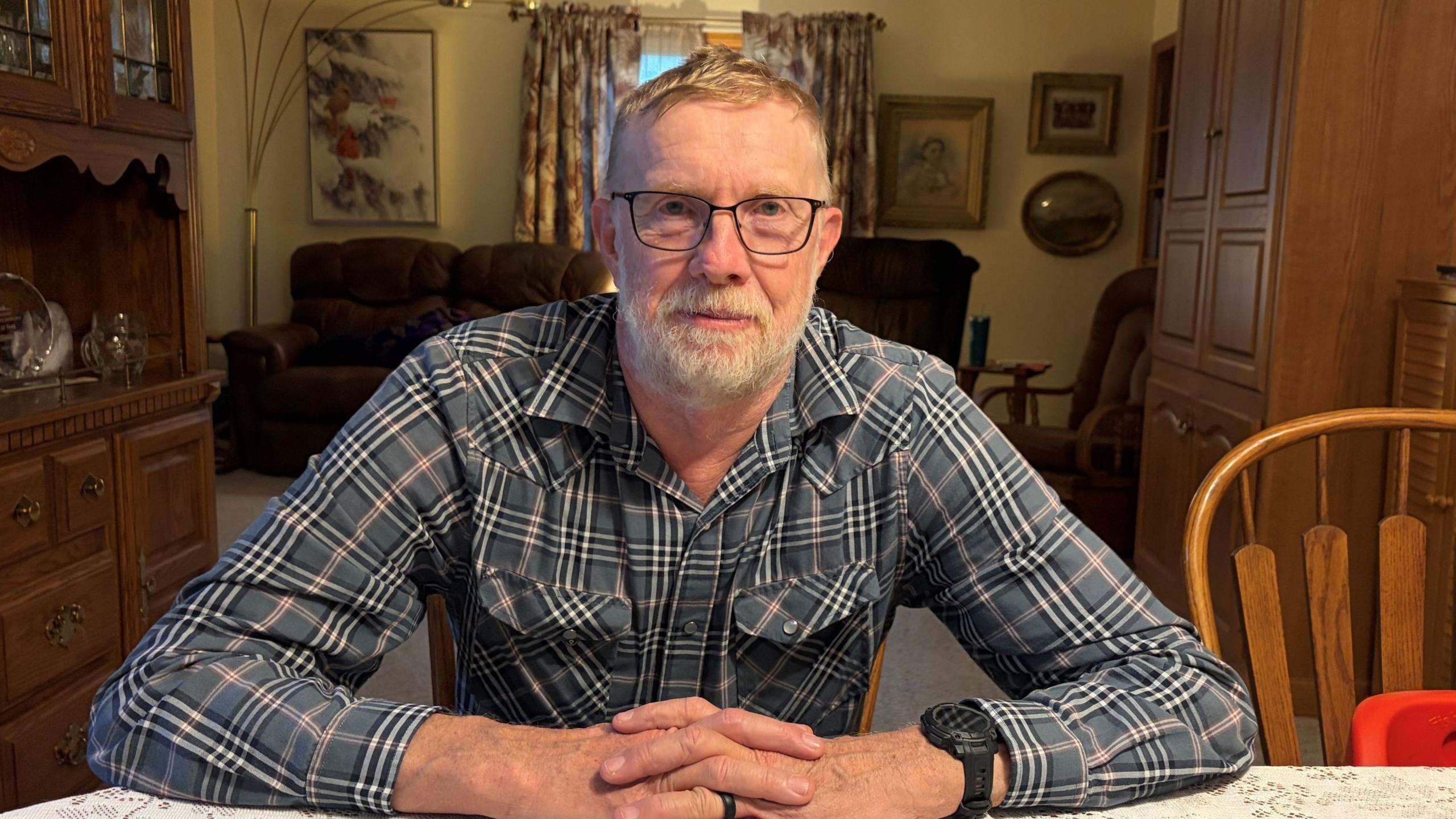 Don Batie wears a plaid shirt and glasses and sits at his kitchen table
