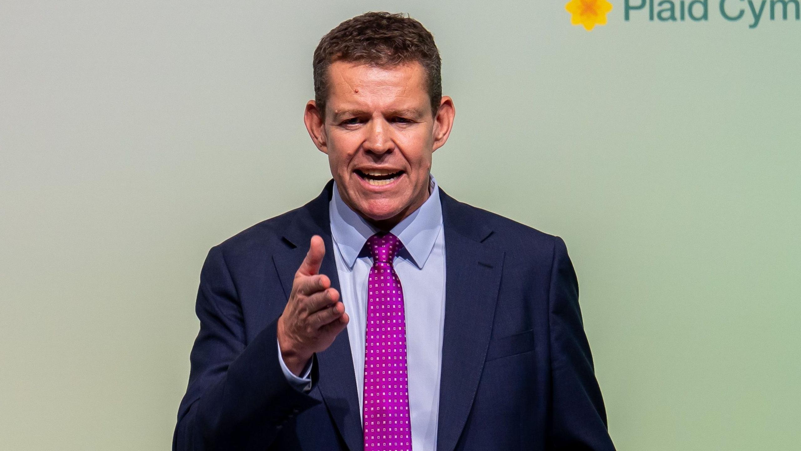 Rhun ap Iorwerth pointing with his right hand. He has a dark blue suit, light blue shirt and purple tie on.