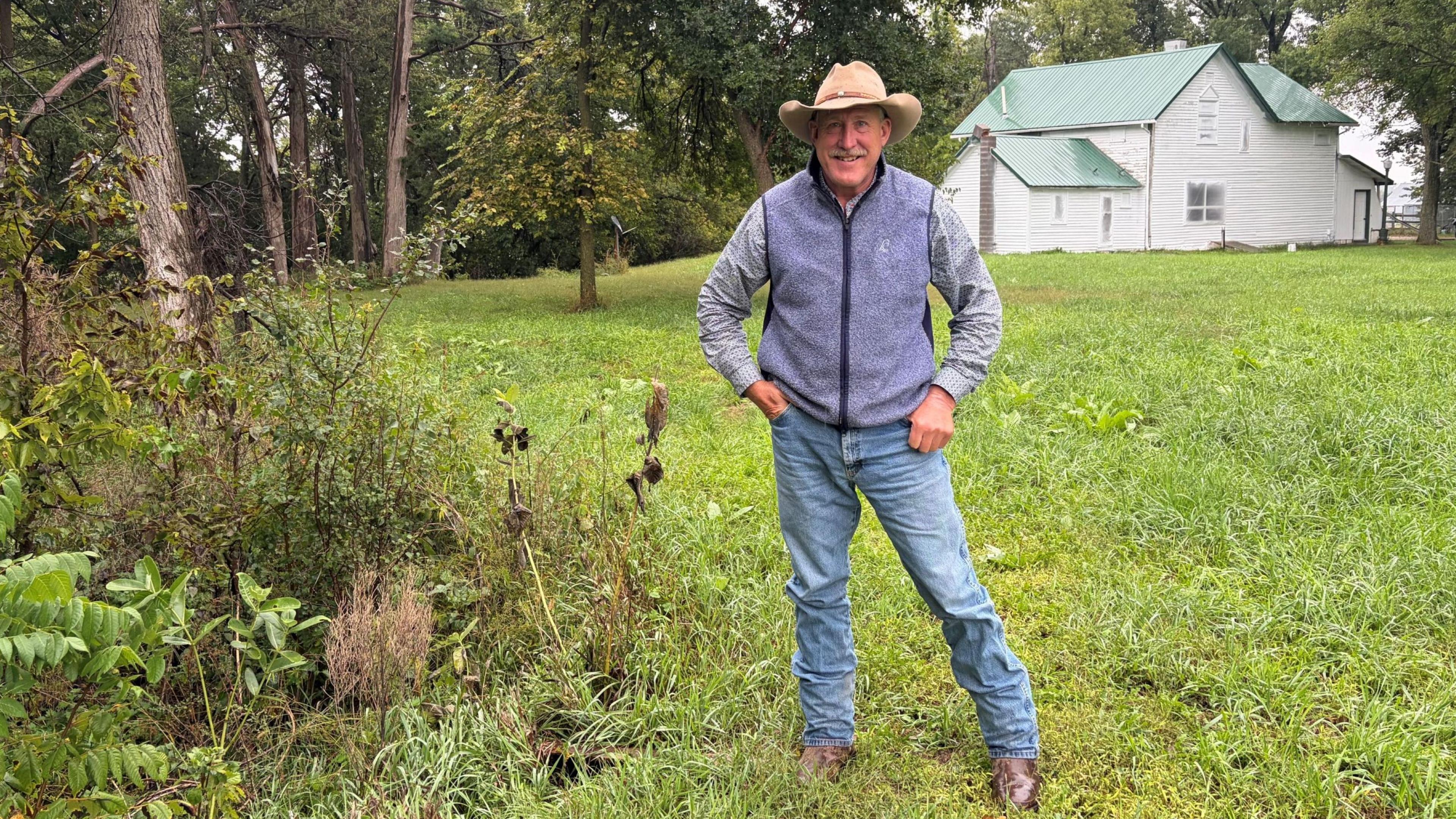 Jim Jenkins, in a cowboy hat, jeans and vest stands on his land
