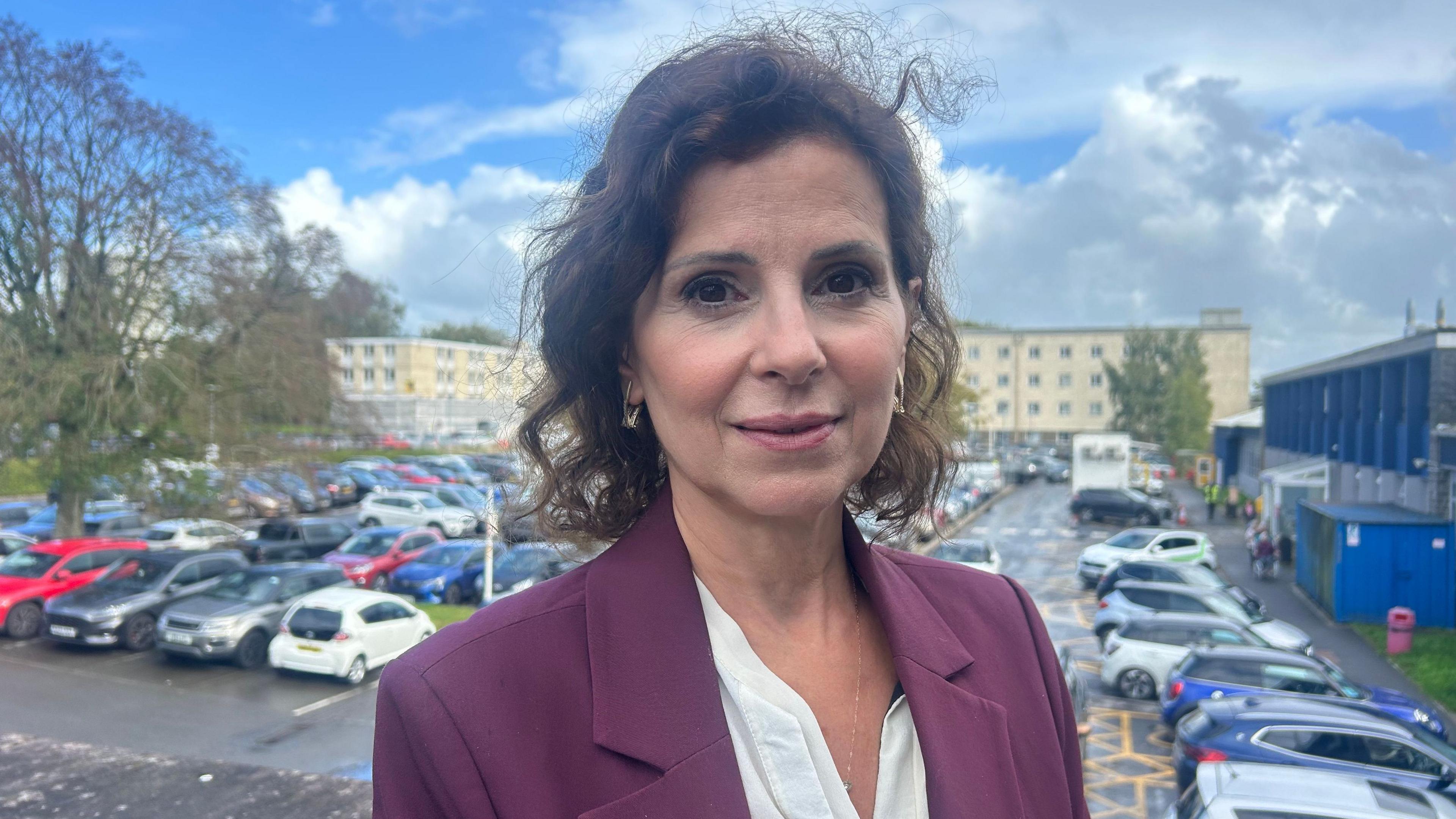 A woman with dark curly hair and a mauve suit jacket over a white shirt is standing in a hospital car park with a large hospital building behind her