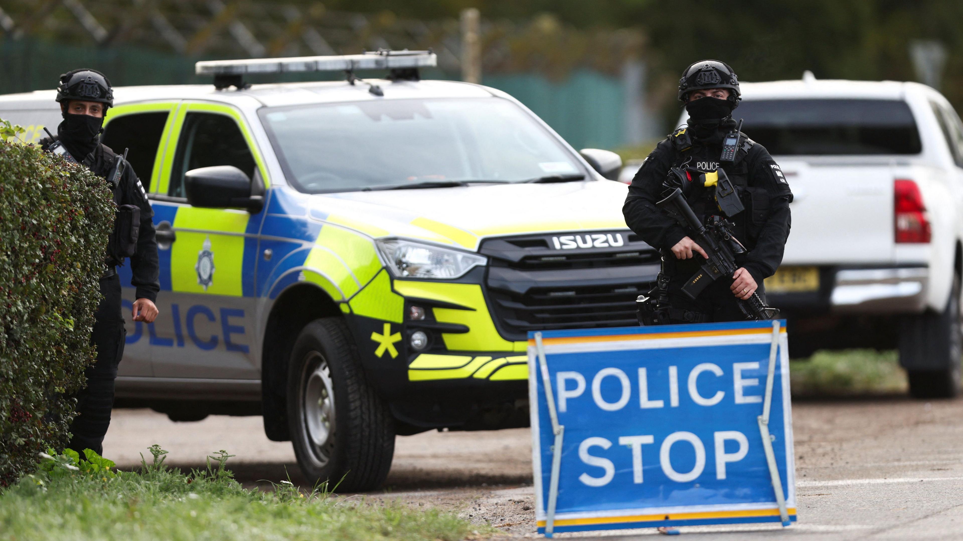 Two police officers clad in black and carrying guns stand in front of a police car, a blue sign reading Police Stop stands in front of one of the officers. Another white truck is parked in the background