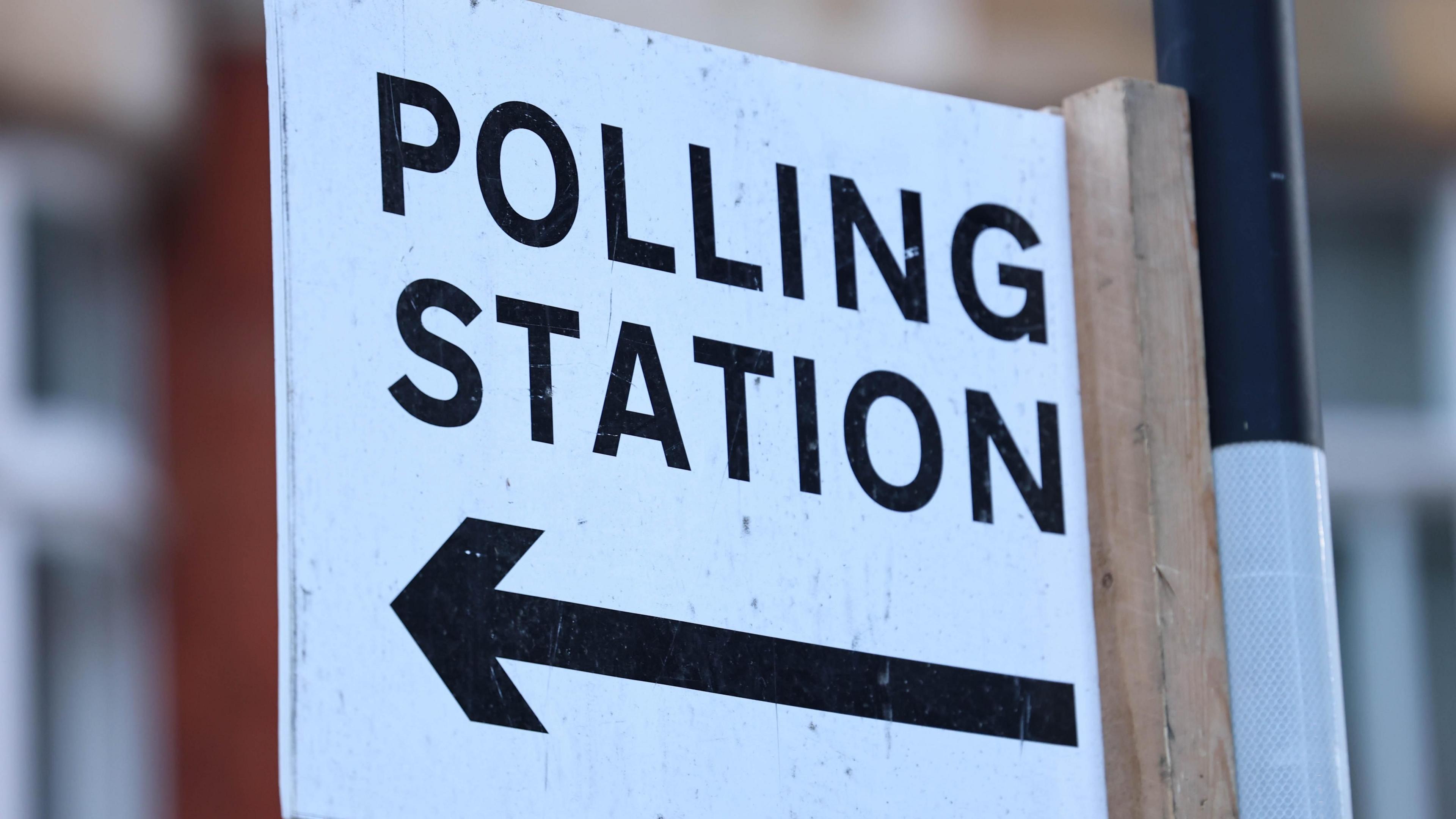 A polling station sign on a post. Polling station is written in black on  white background, with an arrow pointing to the left below the words
