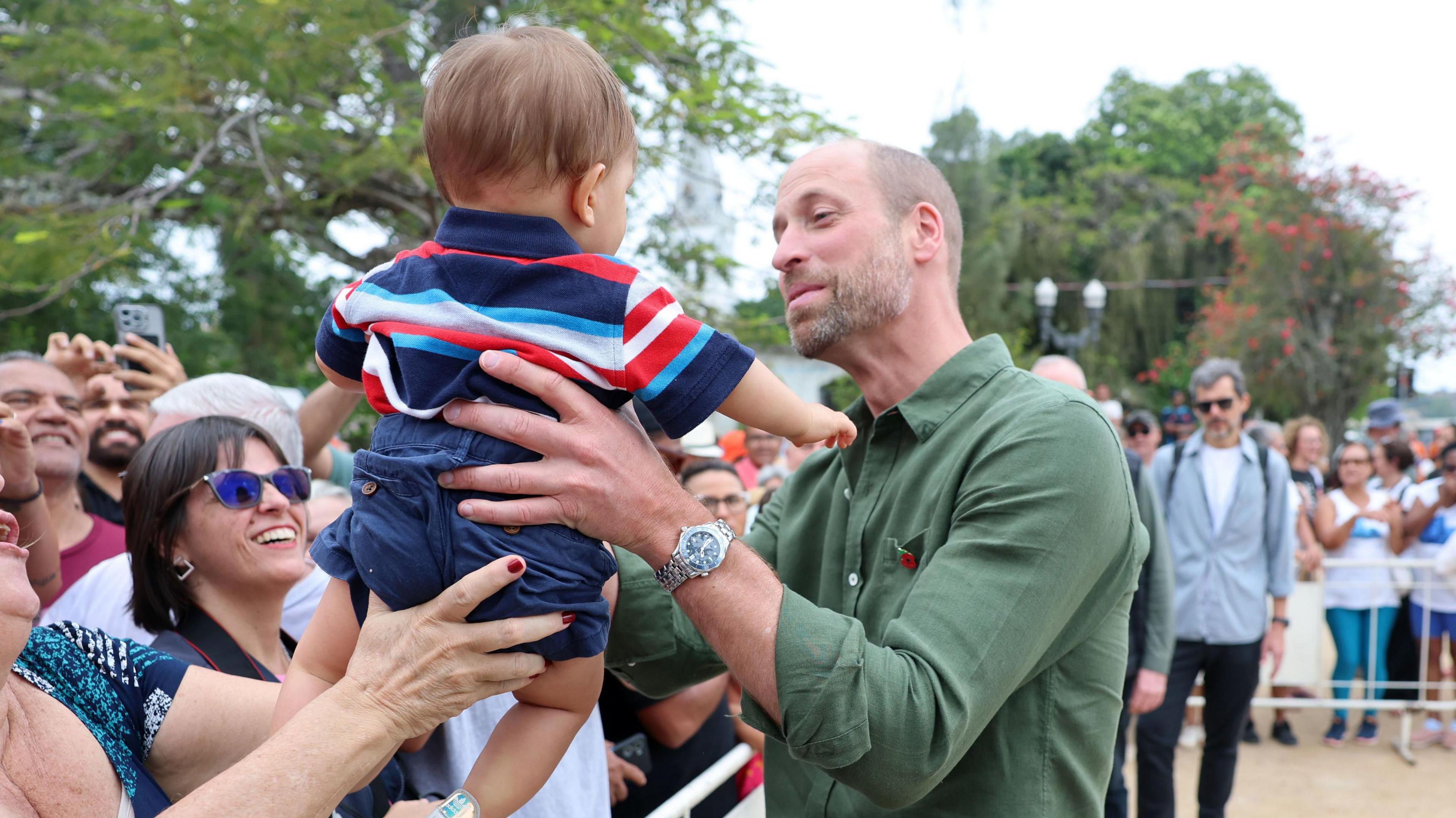 William wearing a khaki shirt with a poppy pin attached to the pocket and he holds a baby boy with both hands. The boy has his back to the camera and is wearing a horizontally striped tshirt in navy, red, turquoise and white, and navy shorts. The boy is being handed to William by a woman who is only in shot by her hands. They are among the crowds of people behind barriers waiting to catch a glimpse of the prince