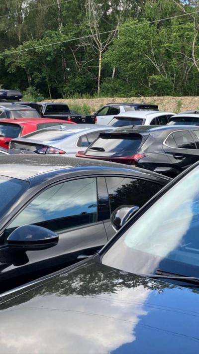 Lots of cars parked closely together with a fence and trees in the background