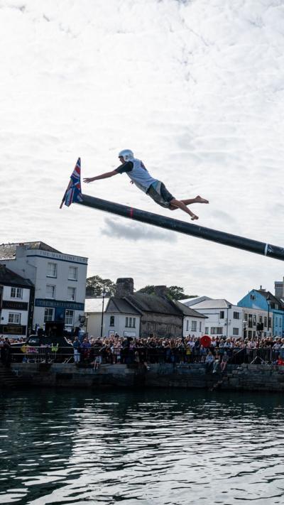 A man leaping for a flag after running up a slippery greased mast. There is a Union Jack flag hanging at the end of it. There are lots of people stood in the background watching on.