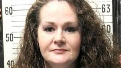 A woman with curly brown hair having a mug shot taken.
