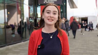 A young woman at Labour conference in Liverpool. She has long auburn hair, and is wearing a navy top and brick-red cardigan.